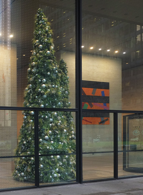 large plainly decorated christmas tree behind a window, orange and black abstract painting on a wall nearby
