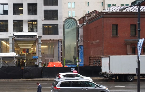 new construction on wellington street, beside an old red brick building, a new curved roof glass atrium, behind black hoardings, and a new entrance