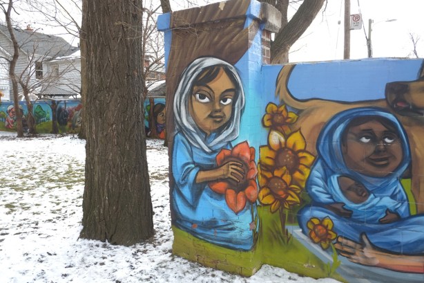 part of a mural along a wall by elicser elliot, a girl with a white head scarf holds a large orange flower. winter time, snow on the ground