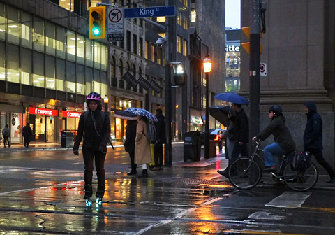 a woman crosses the road, early morning, rain, wet pavement with reflections, she is wearing a bike helmet and roller blades that light up. Pedestrians and cyclists waiting for the light