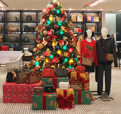 mannequins standing beside a decorated christmas treee in a store, with wrapped presents under the tree, and handbags for sale