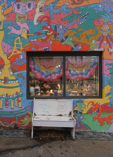 old white bench outside, leaning against wall with window (store) and painted in multicoloured monsters