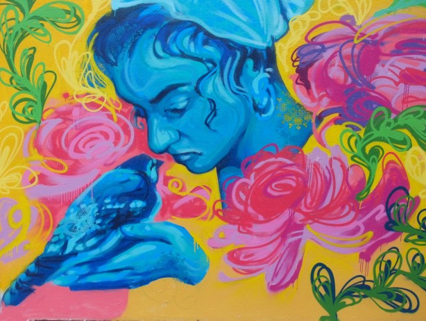 close up view of mural by Amani Muhammad, a woman in blue holds a blue pigeon, pink and red flowers around them as well as blue butterflies