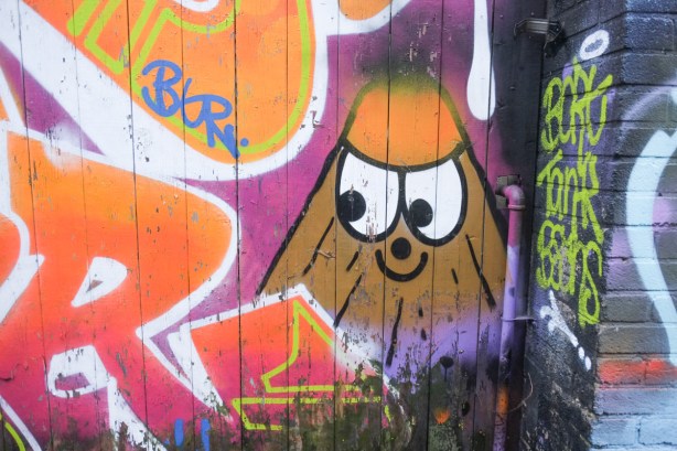 in the corner of a mural, a little brown cone shaped character with big eyes and an orange hat
