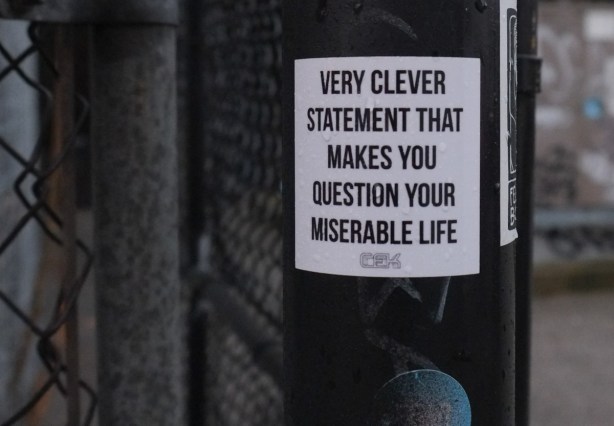 sticker graffiti on a pole, all text, says very clever statement that makes you question your miserable life 