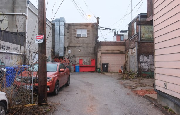 orange car parked in a short alley or driveway, by a pole with a sign that says danger restricted area, backs of buildings, muddy