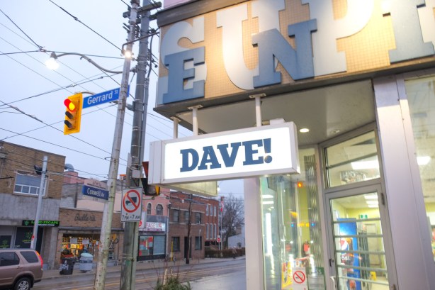 lit sign that says Dave! hanging over the entrance to a convenience store at Gerrard and Coxwell, Coxwell street stores in the background