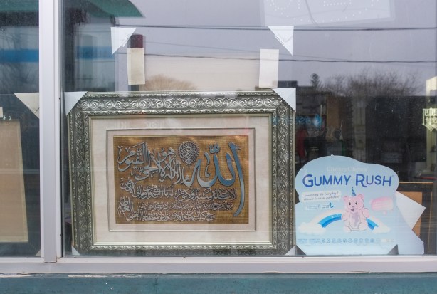 in store window, a framed image of arab calligraphy beside an ad for unicorn shaped gummy rush