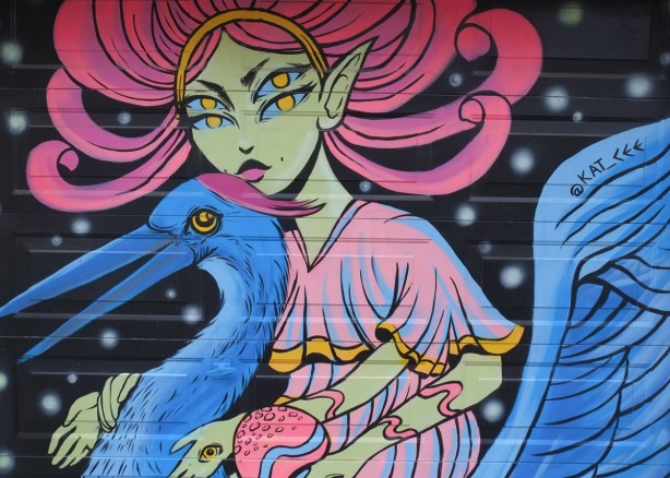 close up of pale green skinned woman in mural of a four-eyed woman with pink flowing hair riding on a blue goose (or pelican), a bird with a long beak. she has an eye on the back of her hand too 
