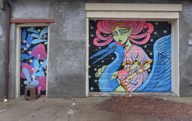two street art paintings on a concrete building, a small painting of pink mushrooms on a door, and a larger mural of a four-eyed woman with pink flowing hair riding on a blue goose (or pelican), a bird with a long beak. she has an eye on the back of her hand too 