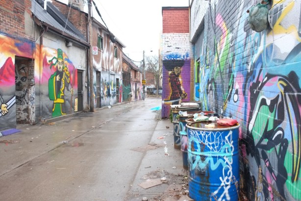 alley with metal oil drums covered with graffiti, low buildings with street art on them, 