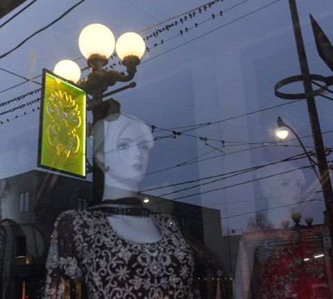 mannequins in store window with reflections of globe shaped street lights and yellow lit public cart banner on light pole