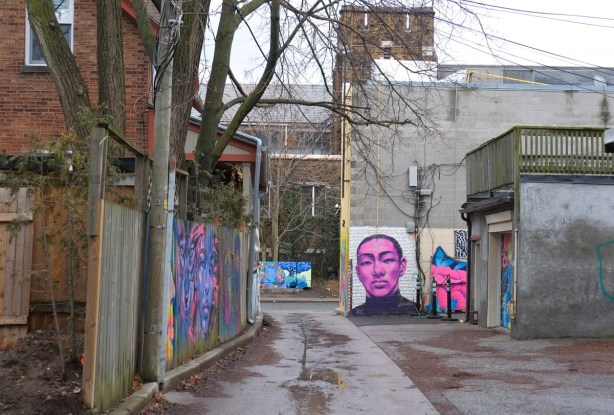 an alley behind the Danforth, a painted fence, a large pink man's portrait, a painted traffic box at the end of the lane