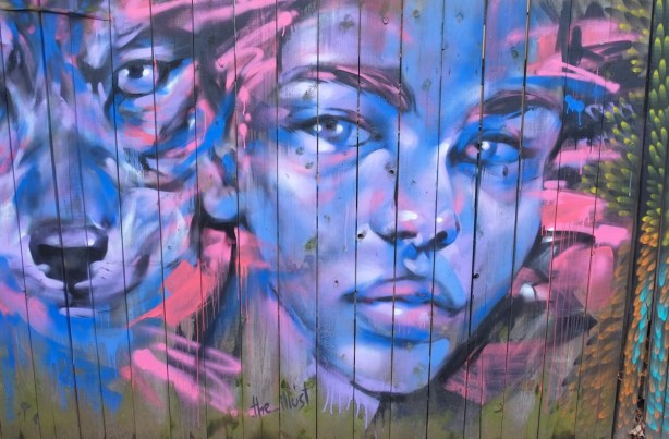 close up of part of a mural on a wood fence in an alley painted by Natsha Dichpan, a purple young woman's face beside a pink and purple wolfs face 