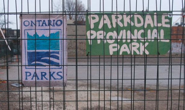 hand painted sign in green and white that say Parkdale Provincial Park, on a metal construction fence around a vacant lot waiting for redevelopment on Brock Ave.., also a hand drawn sign that mimics the blue and white Ontario Parks sign 