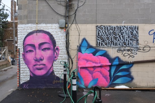 mural on side of concrete building, in two sections, on the left a pink faced man with very short dark hair, surrounded by words, on the right is a square of calligraphy white letters on black with a pink flower below it. The words say sometimes you have to be lion to be the lamb that you are 