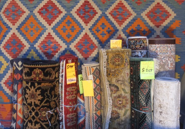 carpets in a store window, some rolled in front of a carpet with diamond shapes in reds and oranges and teal,