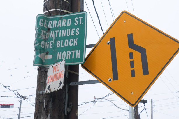 two street signs, a yellow diamond shaped sign that says road narrows and green sign with words Gerrard street continues one block north