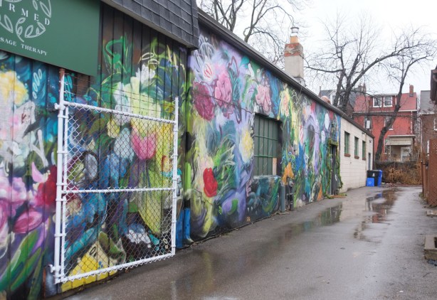 in a driveway or parking lot, with a white metal gate, along one side of a store, a mural of many flowers