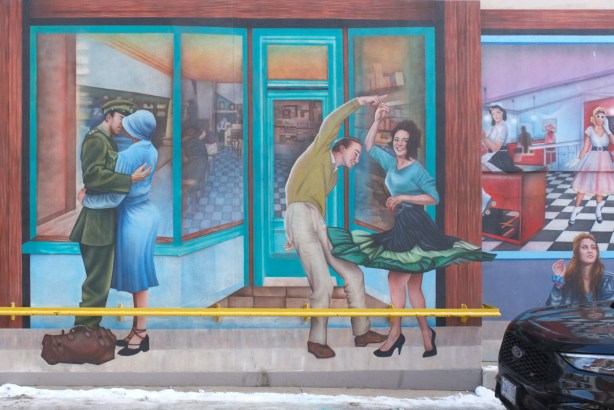part of mural by Christiano De Araujo, windows of time, in Weston, history series, a WW2 couple kissing soldier and woman in blue with blue hat, also a couple dancing from the 1960s. Another window shows interior of soda parlor