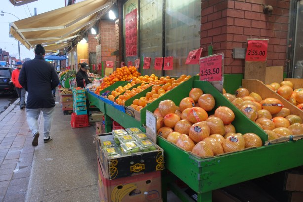 outside display boxes painted green and full of citrus fruits for sale, chinatown store, awning overhead, people walking by 