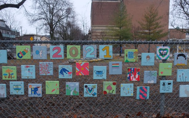 chainlink fence by a school has many little square paintings attached to it, each one was by a student from the class of 2021. 