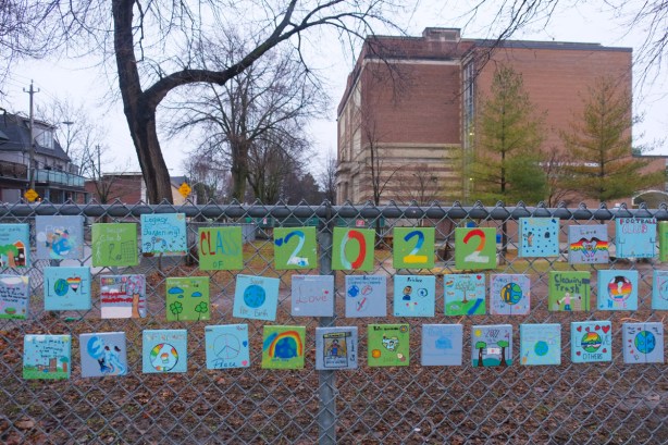 chainlink fence by a school has many little square paintings attached to it, each one was by a student from the class of 2022. 
