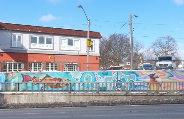 wall beside sidewalk at railway underpass, building in the background, fish, first nations symbols, 