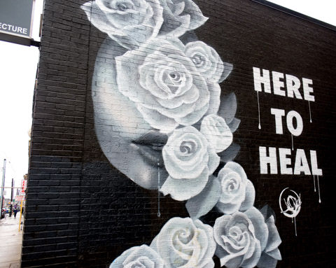large painting in white on black brick white roses, and text that says here to heal, a woman's face is partially hidden by the white roses 