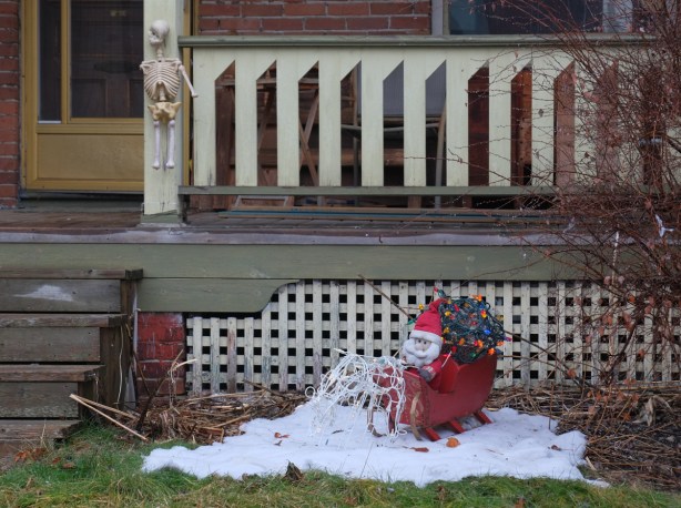a skeleton halloween decoration is on the railing of a porch, in front of it, on the grass is a small santa claus in a red sleigh on a small pile of snow
