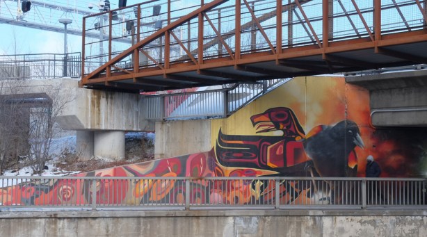large bird like the top of a totem pole, part of a mural, on a wall beside a sidewalk under a railway bridge