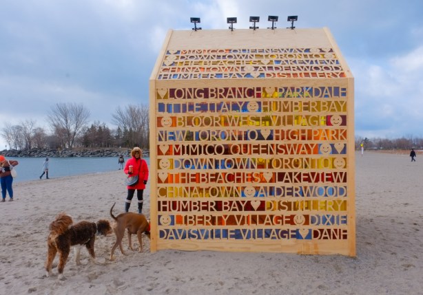 winter stations installation at Woodbine Beach, plywood hut with cut out on the sides spelling out the names of Toronto neighbourhoods. Different colours of plastic covering the names, 