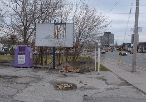purple bin to donate clothing, broken sign, puddle in cracked pavement, street in the background, trees with no leaves