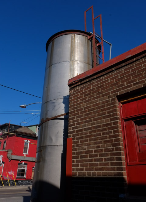 large shiny metal column with a red exterior ladder, beside a brick building with red trim
