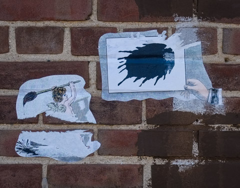 Two paper wheatpaste graffiti on a brick wall. One is a woman with wings on her back holding a very large paint brush over her shoulder, black paint dripping from it. The other is a hand holding a piece of white paper with a very large black ink blot or paint splash on it 