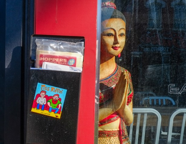 looking in a window at at Thai woman mannequin, wall beside window has a mailbox with a Shoppers drug Mart flyer in it and a sticker on it. 