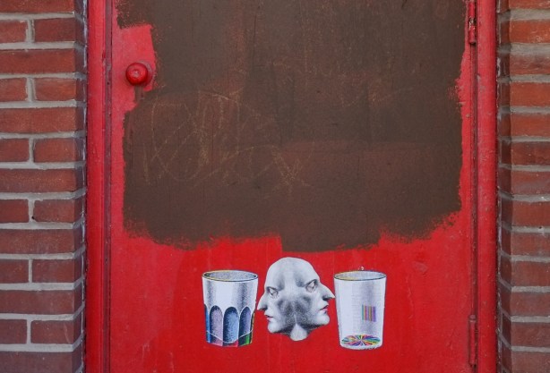three paper graffiti wheatpaste on a red door. in the middle are two identical faces merged together back to back. on the left is a ridged glass drinking glass and on the right is another glass 