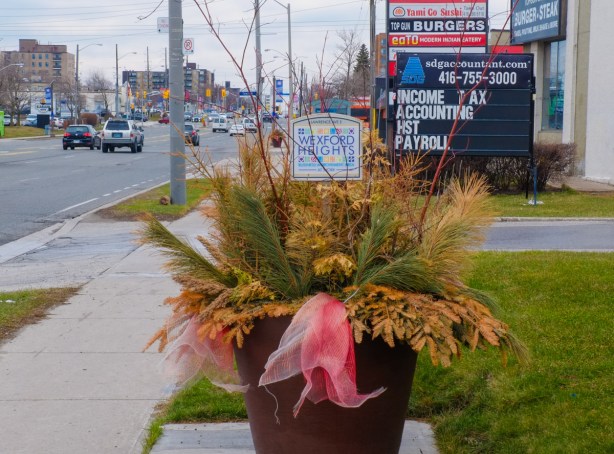 large black planter beside sidewalk with faded red ribbon, winter greenery pine and a wexford heights sign