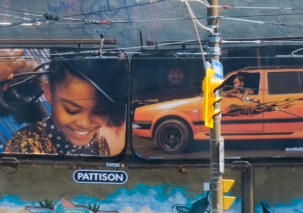 close up of images on billboards. two images. Someone is brushing the hair of a young black woman in one photo. In the other, a person is sitting in a small orange car, viewed from the drivers side of the vehicle. 