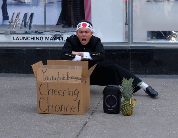 man yelling in Japanese, wearing black outfit and a head band with large red dot on it, beside sign that says 