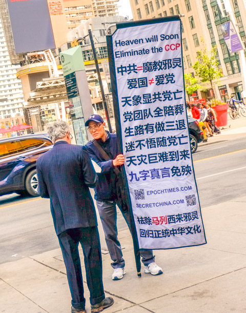 two men talking, one holding a large white banner with blue chinese writing on it.  Some English words, Heaven will soon eliminate the CCP, secret china .com and epoch times, 