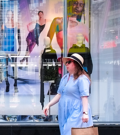 a woman wearing a blue dress and a hat walks past a store window with many reflections