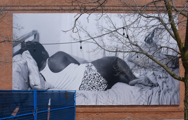 large black and white photograph on an exterior wall, black woman lying on bed with back to the camera, in white t shirt and black and white patterned shorts