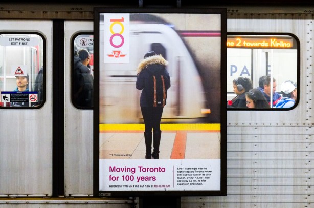 A subway train is stopped behind a poster at the station. on the poster is the back of a woman in a black parka with fur lined hood as she in turn is standing by a ttc subway train