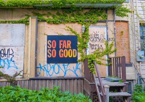 poster on a boarded up window that says so far so good