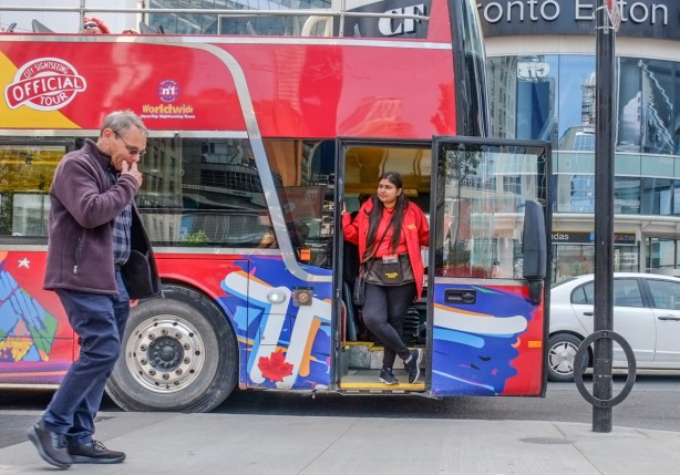 a woman in a red jacket stands in the doorway of a double decker tour bus as a man walks past