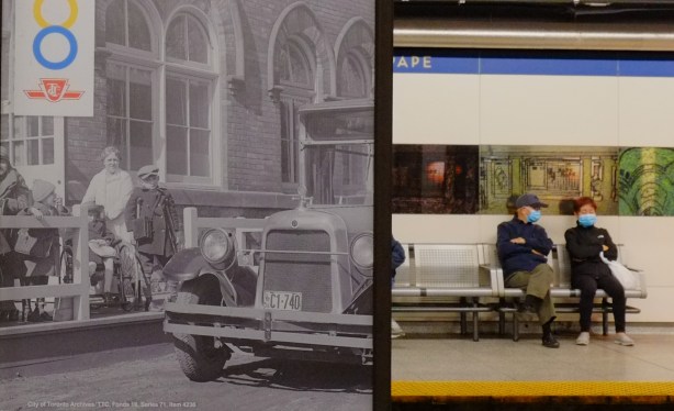 an older Asian couple in covid masks sit on a bench at pape station platform, waiting for a train, posters of black and white old ttc photos on display between the tracks