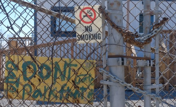 no smoking sign on link fence beside a gate locked with two chains, one rusty and one newer. Also a hand painted sign that says don't park in front of the gate