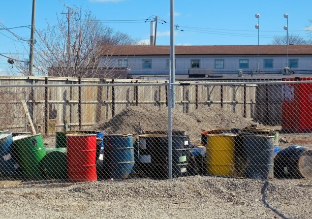 a line of different coloured oil drums along a chainlink fence, wood fence behind