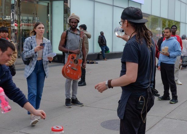 small group watching a street performer at yonge and dundas, one black man is holding a guitar, a woman with a phone, another man putting a coin into a can